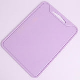 A light purple plastic cutting board with a built-in handle cutout in the top right corner, shown on a solid pale pink background.