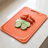 An orange plastic cutting board with a handle, shown on a light beige countertop with sliced cucumbers and tomato wedges on top.