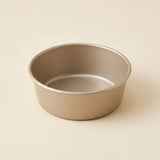 A round metal cake pan with a matte metallic taupe finish, photographed on a clean, solid, warm beige background.