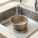 A round beige non-stick baking pan sits in a stainless steel kitchen sink with water running from a chrome faucet.