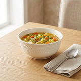 A white ceramic bowl with a textured rim, filled with vegetable soup, set on a light wood table next to a beige napkin and spoon.