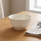 An off-white ceramic bowl with a textured beaded pattern around the upper exterior, shown on a wooden table next to a folded beige cloth napkin and a silver spoon.