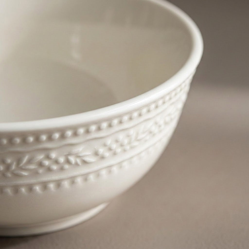 A close-up of a white ceramic bowl with a decorative embossed beaded border and leaf pattern on the exterior, set against a neutral beige background.