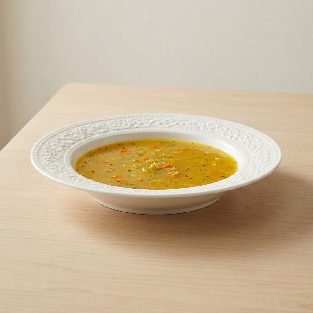 White ceramic soup plate with a wide embossed rim holds golden soup on a light wooden table.