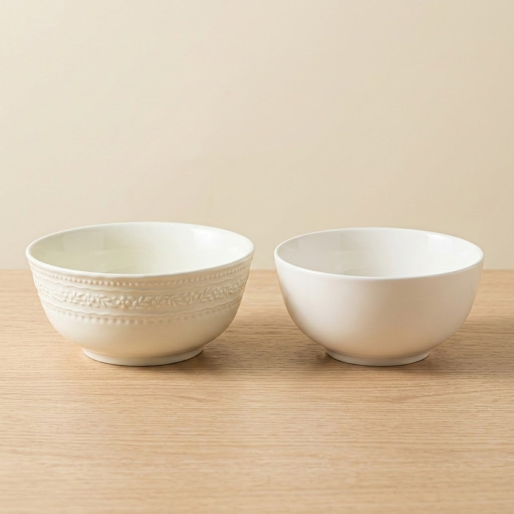Two white ceramic bowls, one with embossed decorative bands and one smooth, sit on a wooden table against a beige background.