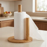 A bamboo paper towel holder with a roll of white paper towels and a sheet draped over the side, set on a kitchen counter with a blurred kitchen background.