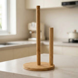 A bamboo kitchen paper towel holder with two upright poles and a round base, shown on a light countertop in a softly blurred kitchen setting.