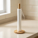 A roll of clear plastic wrap on a wooden stand with a round base, placed on a light speckled countertop in a kitchen setting.