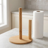 A bamboo paper towel holder with a round base and two upright poles, shown on a light countertop beside a white paper towel roll in a softly lit kitchen setting.