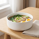 A white ceramic bowl filled with noodle soup, greens, mushrooms, and a halved soft-boiled egg, set on a light wood table next to a folded beige napkin.