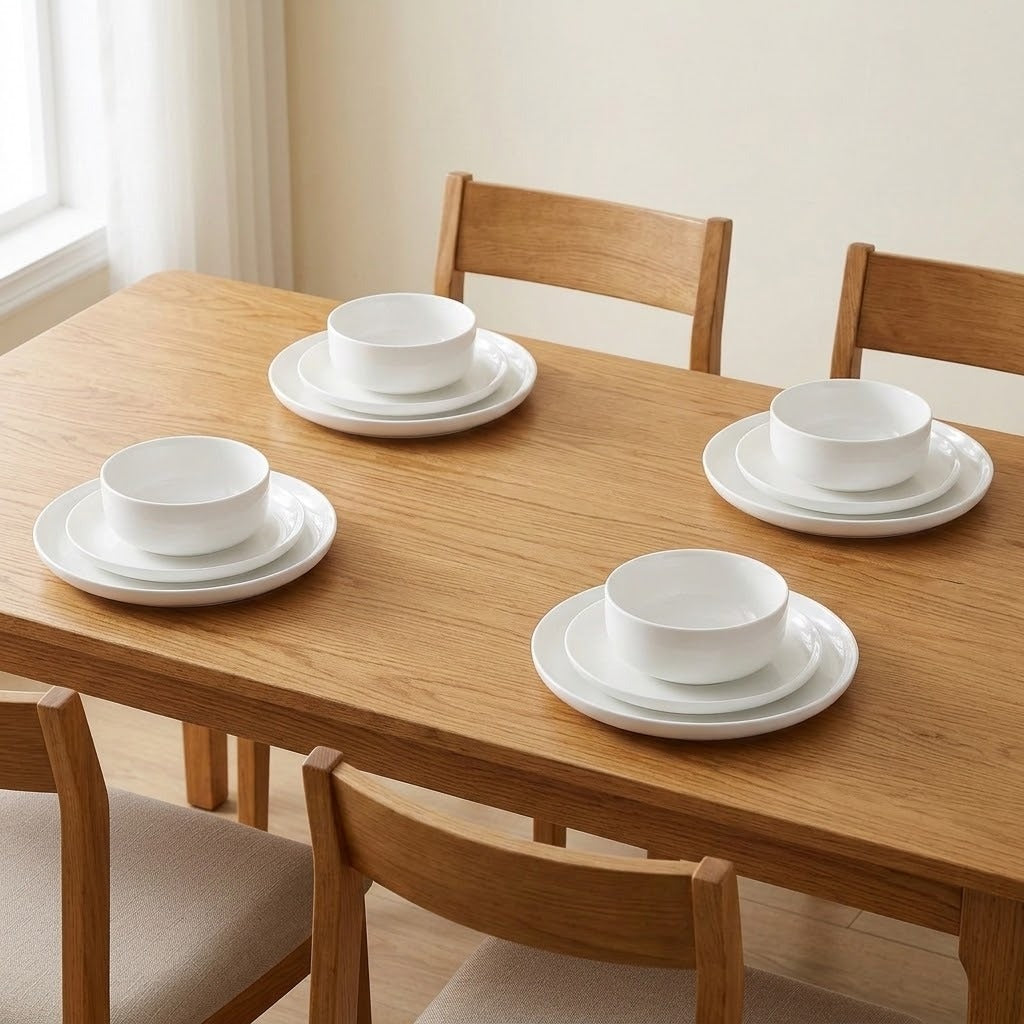 A set of white ceramic dinnerware, including bowls and plates, arranged on a light oak wooden dining table with matching wooden chairs in a bright, minimalist room.