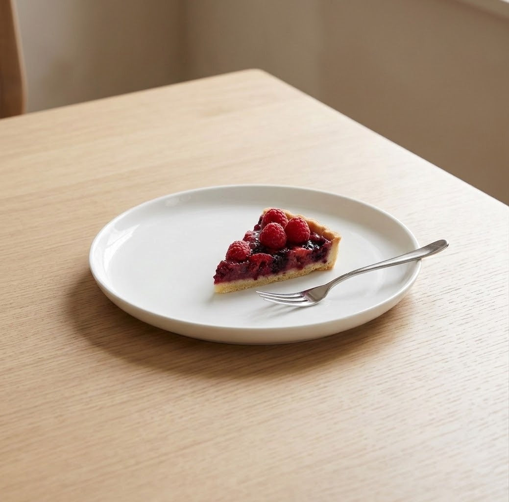 A white ceramic plate on a light wood table, holding a slice of berry pie and a silver fork.