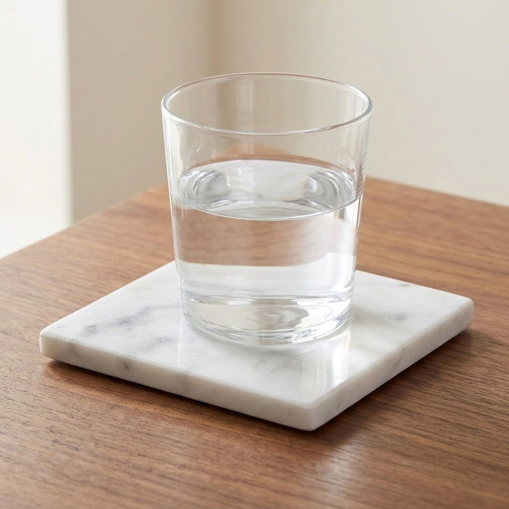 A clear glass tumbler filled with water sits on a square white marble coaster with subtle gray veining, placed on a wooden tabletop.