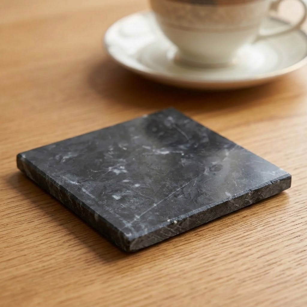 A square dark gray stone coaster with natural white veining, placed on a light brown wooden table with a blurred white teacup and saucer in the background.