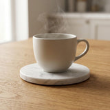 A matte off-white ceramic coffee cup with steam rising from it, sitting on a round white marble coaster on a wooden table.
