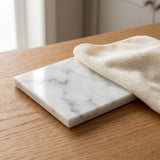 A square white marble tile coaster with gray veining, resting on a light brown wooden table with a cream-colored textured cloth draped over it.