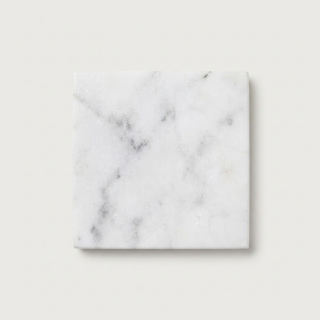 A square white marble coaster with natural gray veining, displayed on a clean, light beige background.