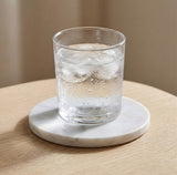 A clear glass tumbler with condensation, filled with a clear iced drink, resting on a round white marble coaster on a light wood table.