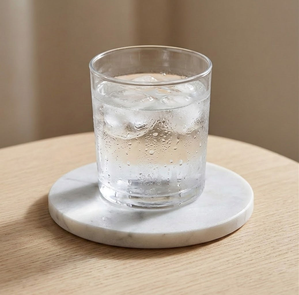 A clear glass tumbler with condensation, filled with a clear iced drink, resting on a round white marble coaster on a light wood table.