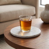 A clear lowball glass filled with amber liquid sits on a round white marble coaster placed on a round wooden table in a softly lit living room.