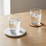 Two clear drinking glasses filled with water and ice, resting on round coasters on a light wood table. One coaster is white marble and the other is natural wood.