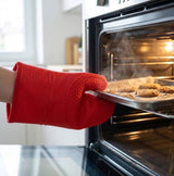 A red silicone oven mitt with a honeycomb texture is being used to remove a baking tray of cookies from an oven in a kitchen setting.