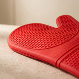A close-up of a bright red silicone oven mitt with a raised honeycomb texture, resting on a light beige stone surface.