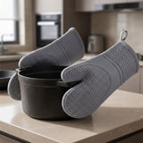 A pair of gray silicone oven mitts with a honeycomb textured grip, shown resting on a black pot on a kitchen countertop with a blurred kitchen background.