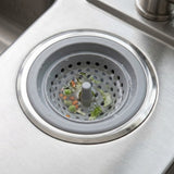 A gray silicone kitchen sink drain strainer basket with a central plug, shown inside a stainless steel sink basin with food debris visible inside.