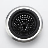 A top-down view of a kitchen sink drain assembly with a brushed silver metal rim, a black rubber gasket, and a black perforated basket strainer in the center, set against a plain light gray background.