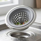 A gray silicone and stainless steel kitchen sink drain strainer basket shown floating above a wet sink, with small food pieces visible inside.