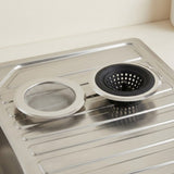 A stainless steel kitchen sink drain assembly with a mesh strainer basket and a black rubber drain basket, resting on a matching stainless steel sink drain board.