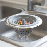 A gray and silver kitchen sink drain strainer basket with a vegetable basket insert, shown in a stainless steel sink with water running under it.