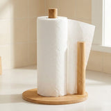 A bamboo paper towel holder with a roll of white paper towels and a sheet extending from the side, set on a light countertop against a beige tiled kitchen background.