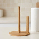 A bamboo paper towel holder with a round base and two vertical stand arms, shown on a light countertop next to a roll of paper towels.