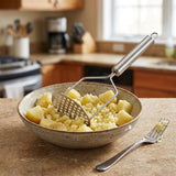 A stainless steel potato masher with a long handle resting in a speckled beige ceramic bowl filled with mashed potatoes, set on a kitchen counter with a fork nearby.