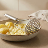 A silver metal potato masher rests across whole and mashed yellow potatoes in a shallow ceramic bowl on a beige countertop.
