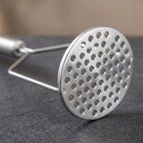 Close-up of a silver metal potato masher with a round perforated plate resting on a dark grey textured surface.