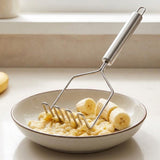 A stainless steel potato masher with a long handle resting over a ceramic bowl containing mashed banana and banana slices, set on a light countertop near a window.