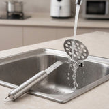 A stainless steel kitchen utensil with a long handle and perforated round head resting in a sink under running water, with a blurred kitchen background.