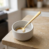 A small speckled ceramic bowl filled with coarse salt, holding a gold spoon, set on a beige stone countertop in a softly blurred kitchen setting.