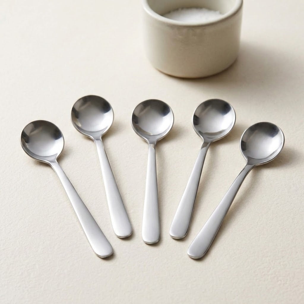 Five stainless steel spoons arranged in a fan shape on a light beige textured surface, with a small cream-colored ceramic cup partially visible in the background.
