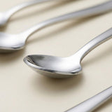 A close-up of polished stainless steel spoons on a warm beige background, with one spoon in sharp focus and others softly blurred in the background.