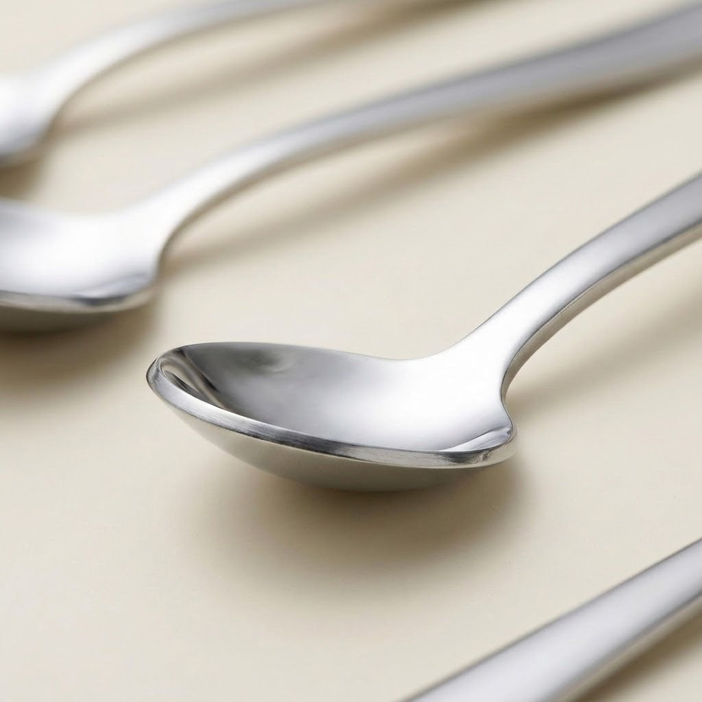 A close-up of polished stainless steel spoons on a warm beige background, with one spoon in sharp focus and others softly blurred in the background.