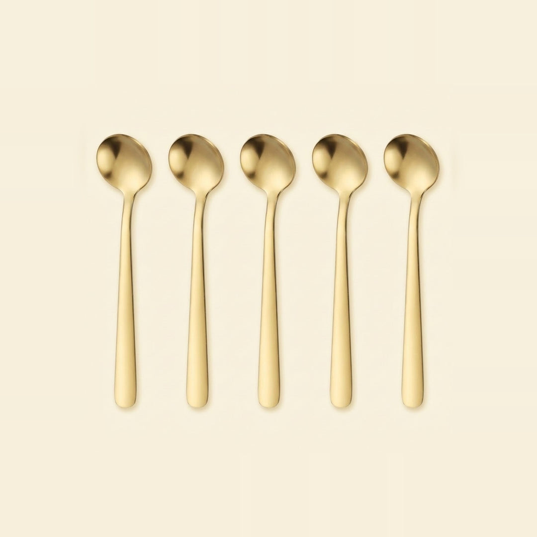 Five gold-colored metal spoons arranged in a row against a solid cream-colored background.