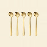 Five gold-colored metal spoons arranged in a row against a solid cream-colored background.