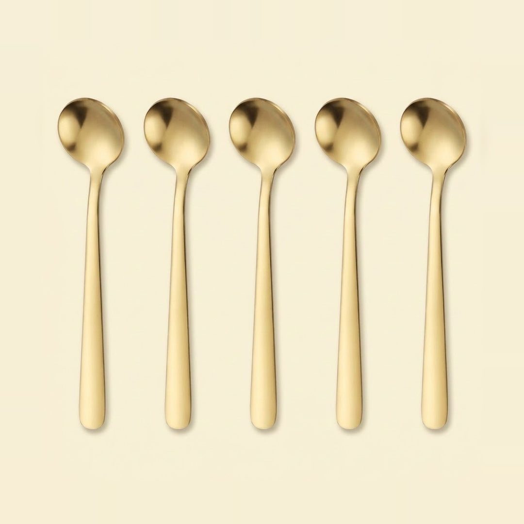 Five slim gold-toned metal spoons arranged upright in a row on a plain warm cream background.