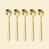 Five slim gold-toned metal spoons arranged upright in a row on a plain warm cream background.