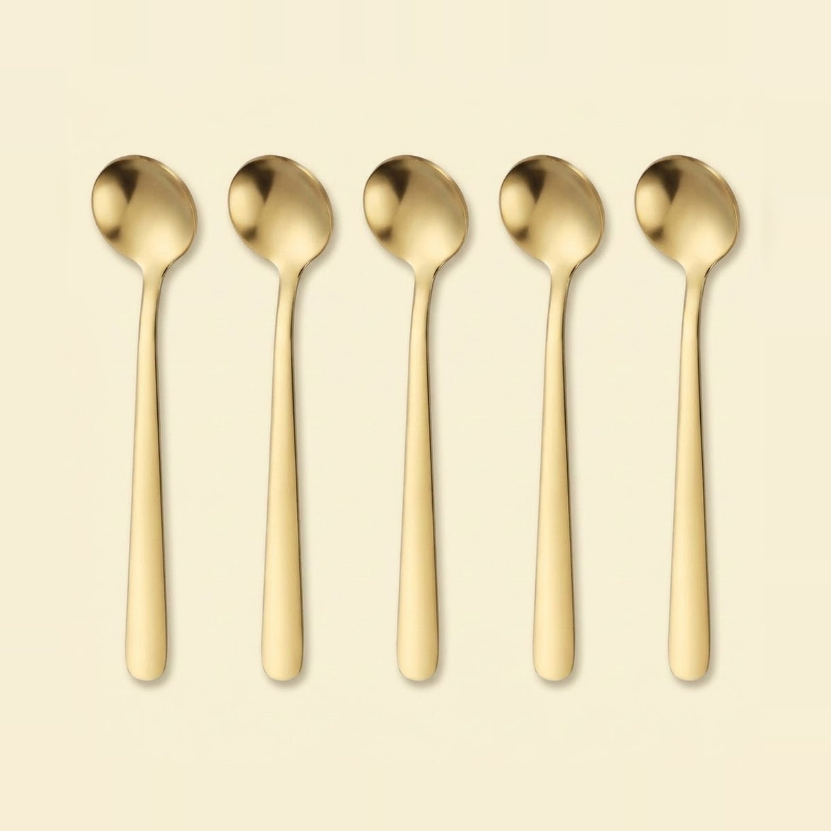 Five slim gold-toned metal spoons arranged upright in a row on a plain warm cream background.