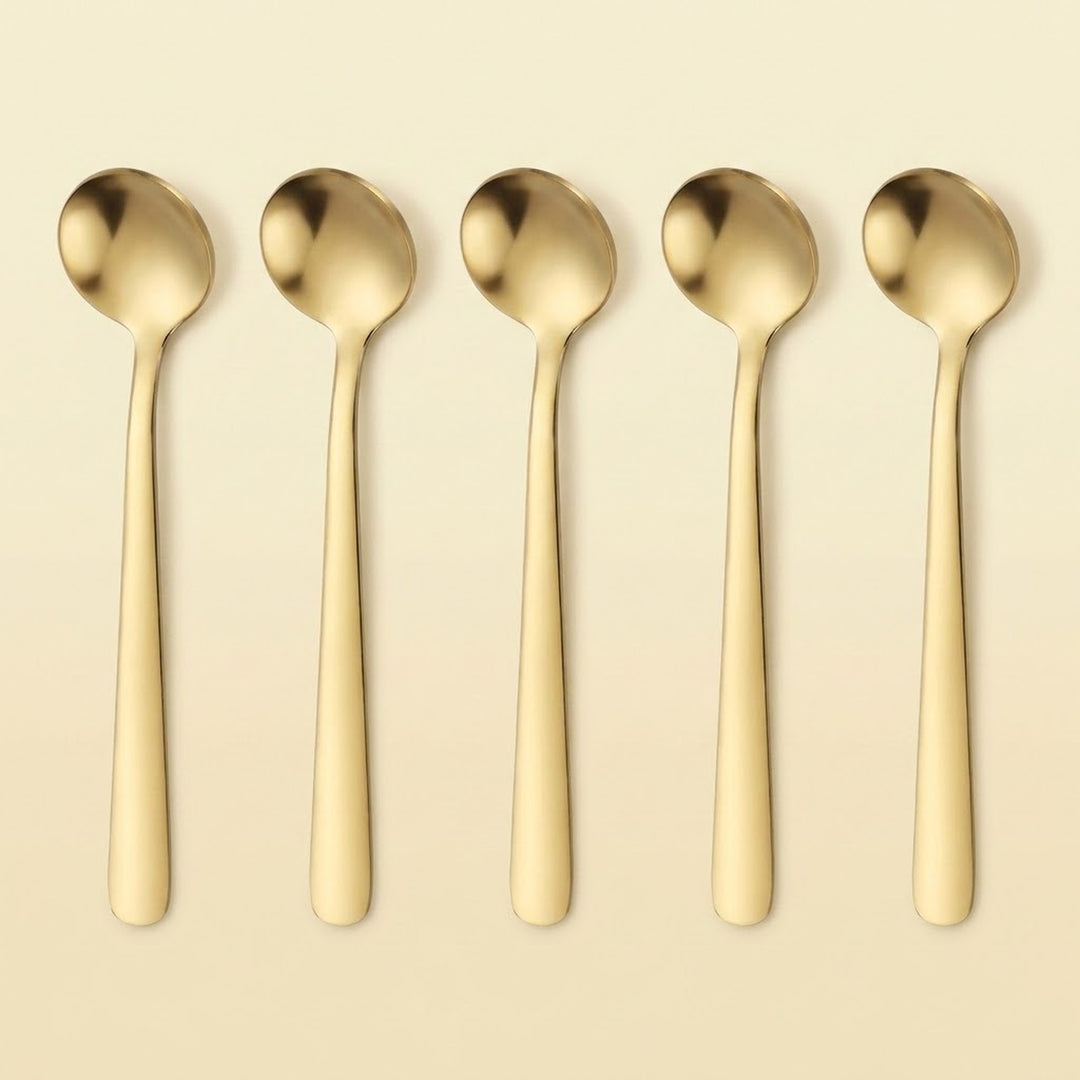 Five gold-toned metal spoons arranged in a row on a light beige background.